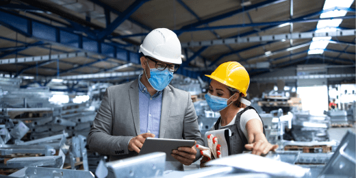 Two workers wearing face masks and following safety protocols, reviewing information on a tablet inside a warehouse.