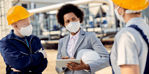 An HR health and safety manager in a grey suit and face mask uses a tablet to discuss protocols with construction workers at an industrial site.